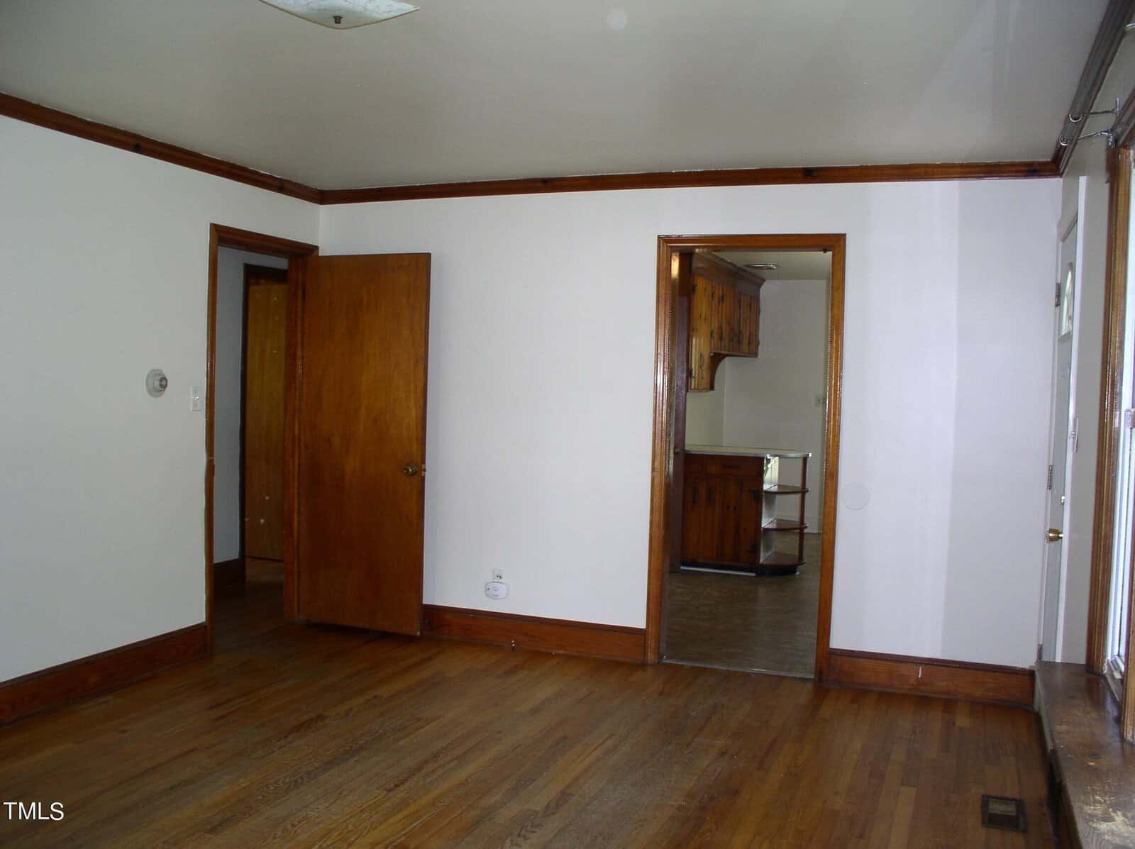 A vacant room with light walls and a wooden floor. Two open wooden doors lead into adjacent rooms, one showing a glimpse of a kitchen with a dining area. Moldings match the wood of the doors. Large windows and a light fixture are partially visible.