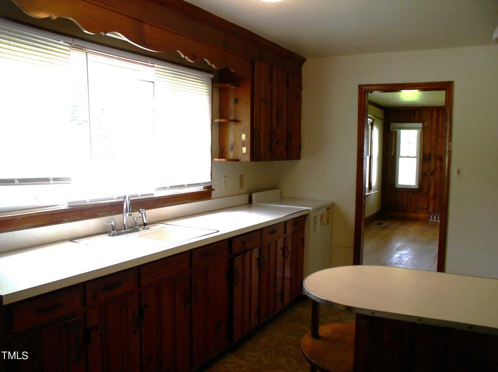 A kitchen with wooden cabinets, a large window above the sink, and white countertops. There's an open doorway leading into another room with wood paneling and large windows. A small round table is positioned near the cabinets on the right.