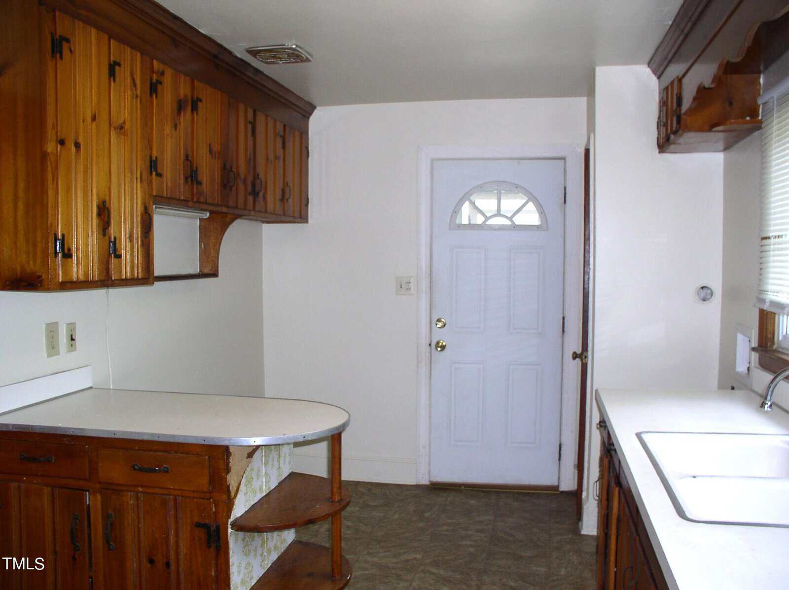 A white kitchen with wooden cabinets, a white countertop, a sink, and a white door with a window. There is a curved countertop with open shelving underneath. Tan floor tiles cover the kitchen floor.