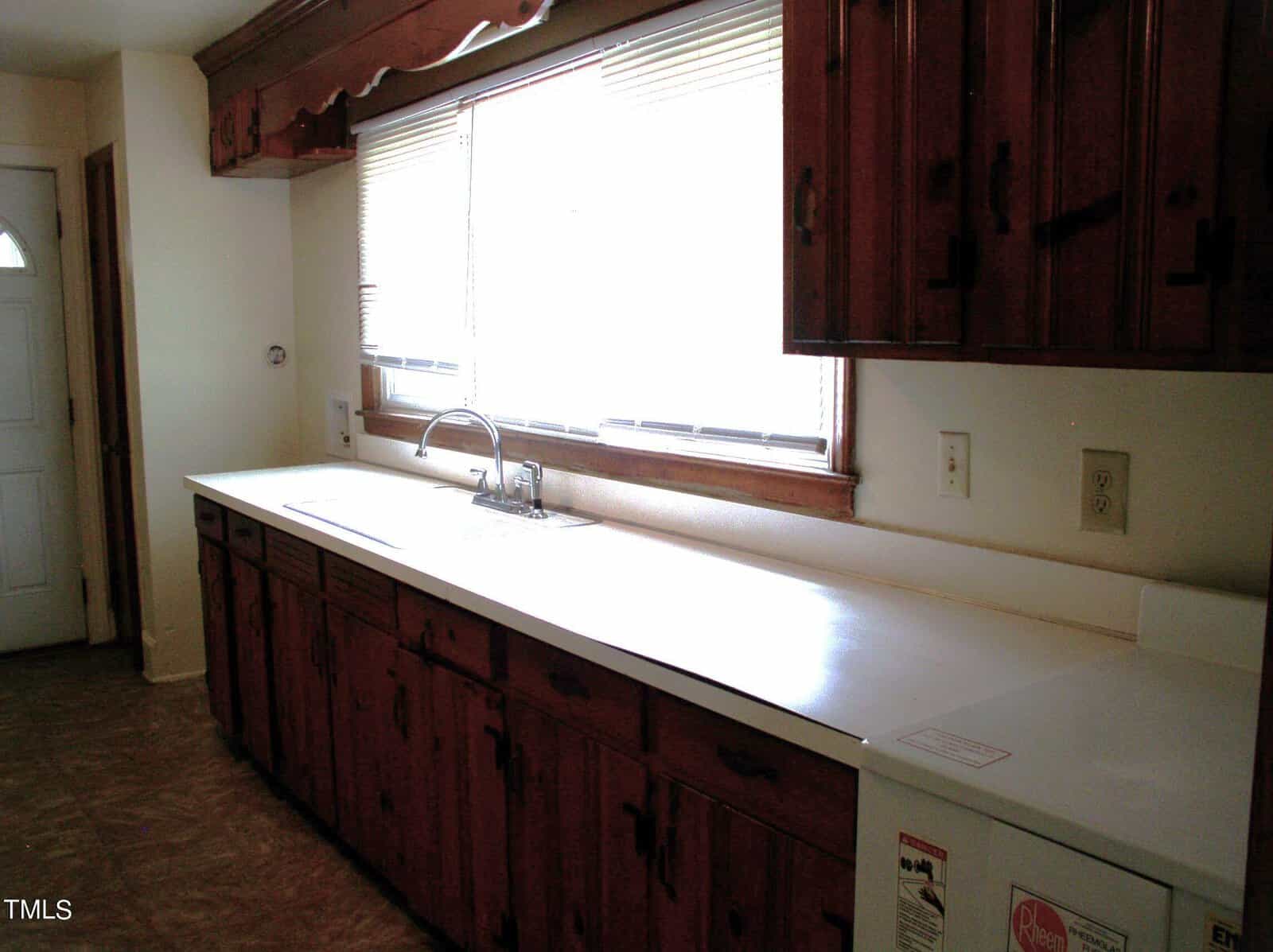 A kitchen with dark wooden cabinets, a long white countertop, and a stainless steel sink beneath a window. The floor is covered in tiled linoleum. A white door is visible to the left side of the image.