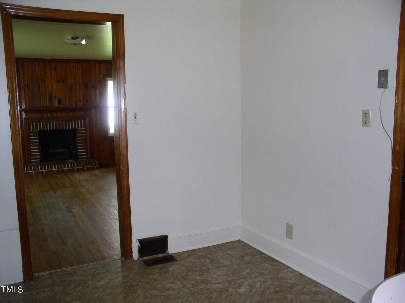 A small empty room with white walls and a beige tiled floor. An open doorway with wooden trim reveals an adjacent room with wooden paneling, a brick fireplace, and a wooden floor. There are light switches on the wall. Natural light enters through the distant window.