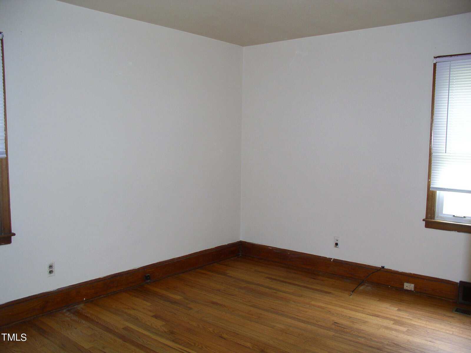 An empty room with hardwood flooring, white walls, and a single window with partially opened blinds. Electrical outlets and cables are visible near the baseboards. The room has minimal decor and is well-lit by natural light coming through the window.