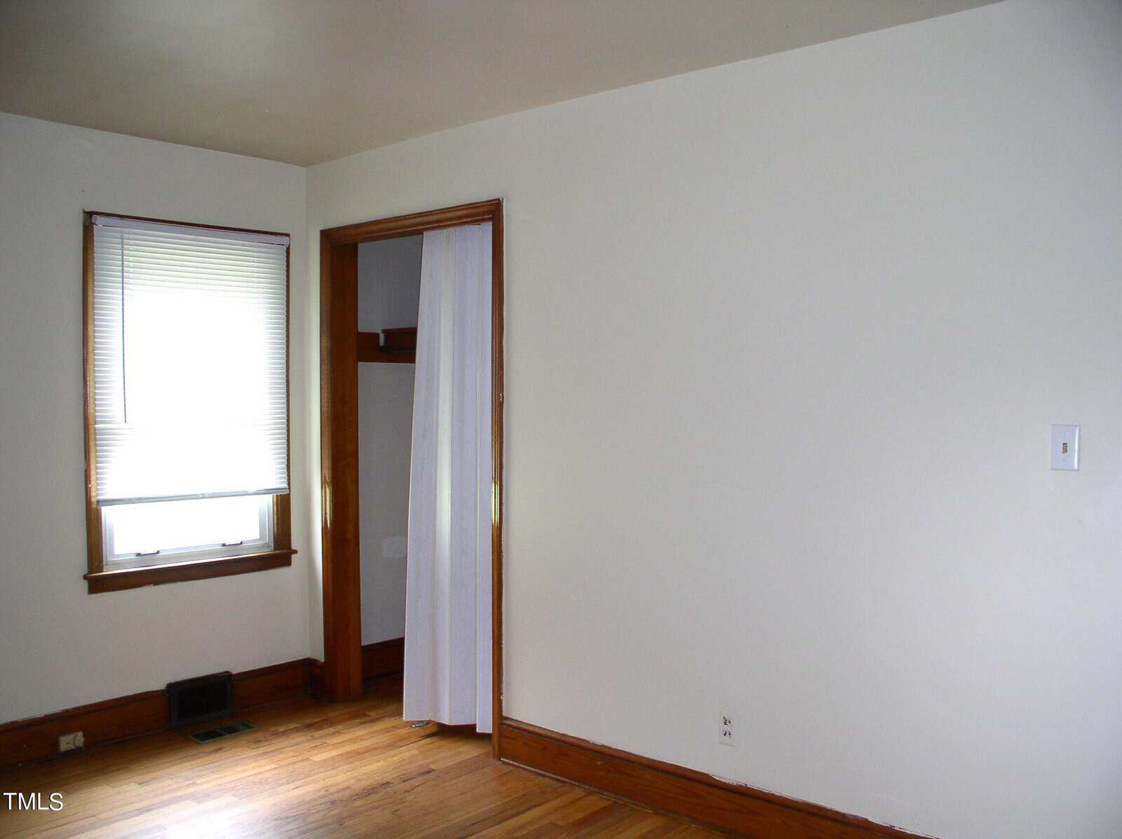 A simple room with white walls, hardwood floors, and a single window with blinds. There is an open closet with sliding doors, one of which has a sheer white curtain. A light switch is visible on the right side of the wall.
