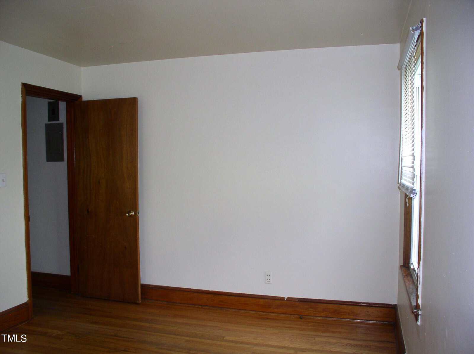 An empty room with white walls, a wooden floor, a brown wooden door partially open, and a window on the right side with blinds partially open.