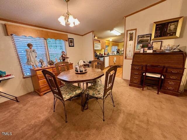 A cozy dining area with a round wooden table and four metal chairs with patterned cushions. The room is adorned with a chandelier, decorations on the dresser by the window, and family photos on the wall and above a wooden writing desk. The kitchen is visible in the background.