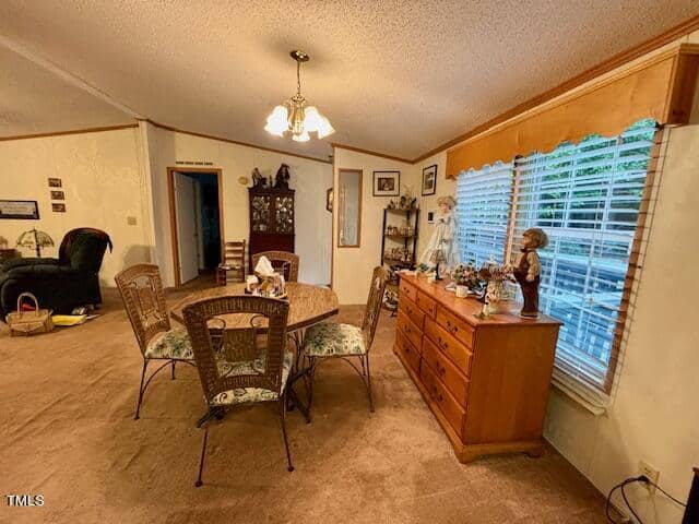A cozy dining room with a round table and four woven chairs. A wooden sideboard with decorations sits by the large window to the right, which is draped with blinds. The room features carpet flooring, light-colored walls, and a chandelier above the table.