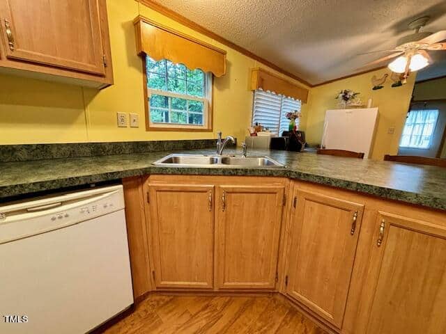 A kitchen with light wood cabinets and green countertops. It features a double sink beneath a window with yellow curtains. There is a dishwasher on the left, a ceiling fan above, and a white refrigerator beside a small window with white blinds.