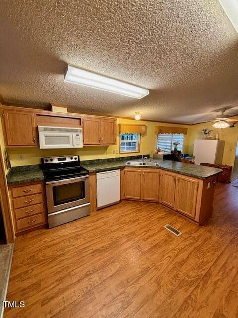 A kitchen with light wood cabinets and green countertops. It features a stove, a microwave, and a dishwasher. The walls are painted yellow, and the floor is wood. The ceiling has a fluorescent light fixture. A large window and ceiling fan are visible in the background.