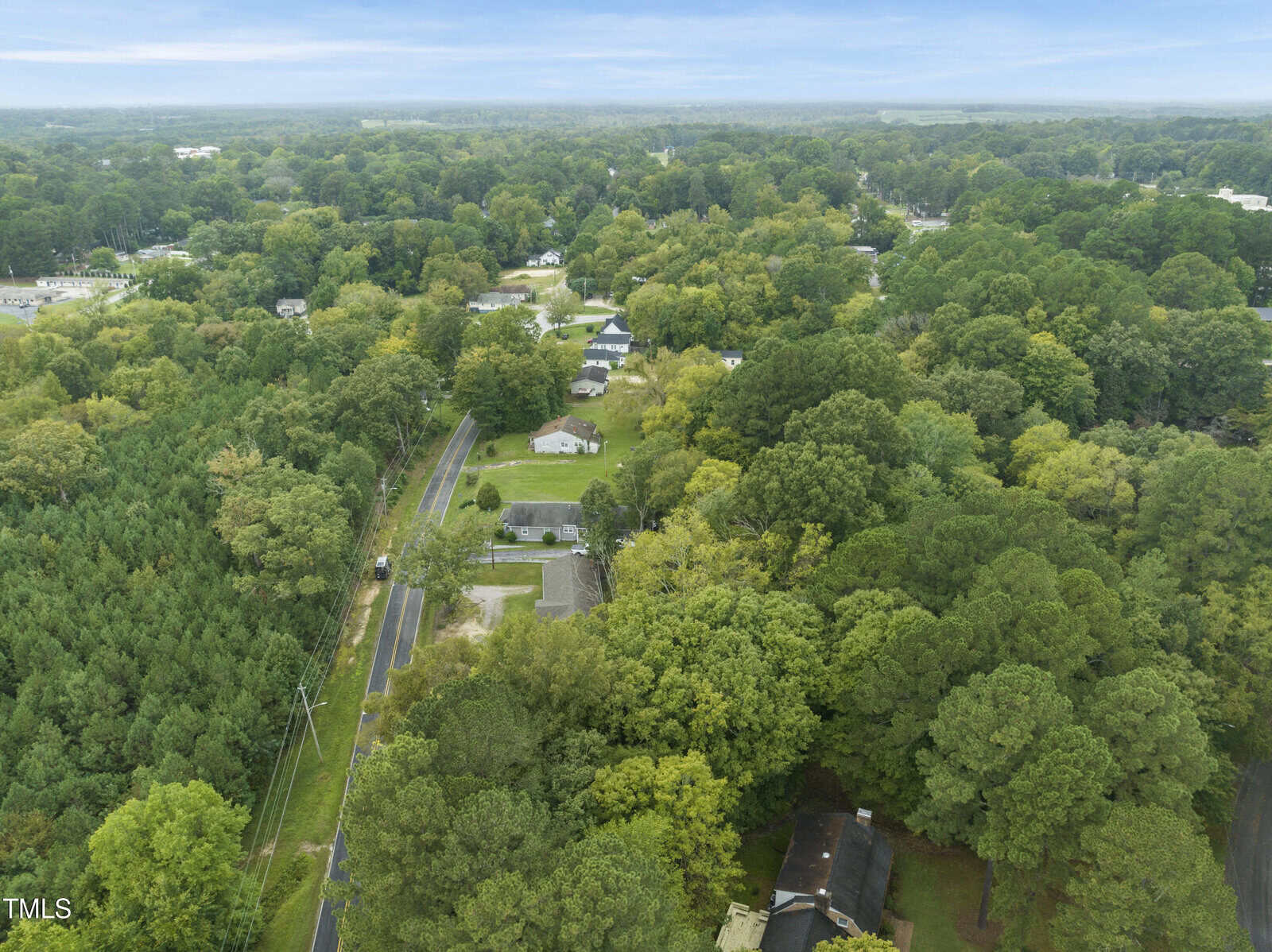 Aerial view of a suburban area surrounded by dense green trees, with houses scattered along a winding road and a partly cloudy sky overhead.