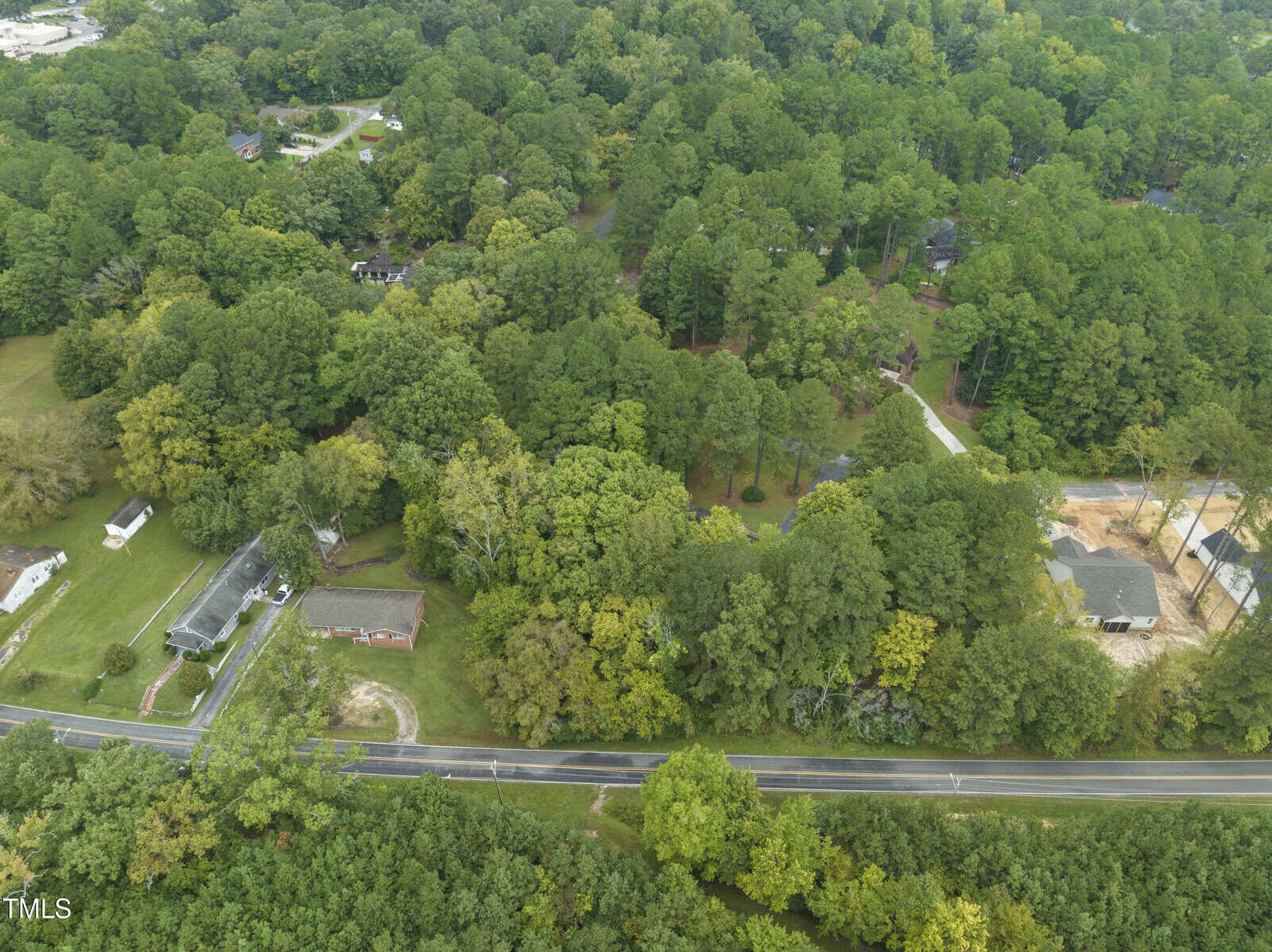 Aerial view of a suburban area with houses surrounded by dense green trees. A two-lane road runs horizontally across the bottom of the image, with homes, driveways, and open yards visible nearby.
