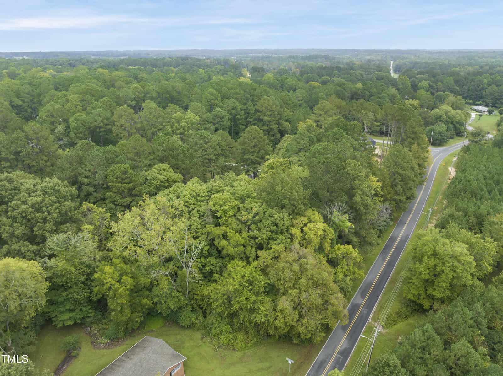 A winding two-lane road bordered by dense green trees and forested areas, stretching into the distance under a cloudy sky. Part of a house roof is visible in the lower left corner.