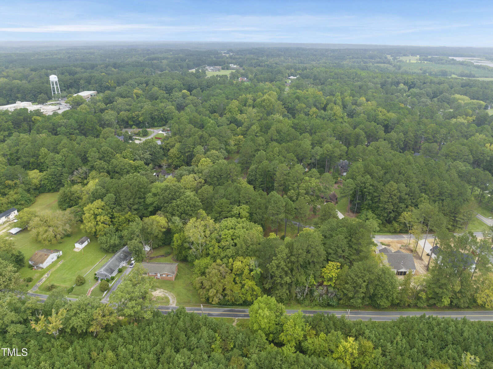 Aerial view of a rural area with dense green trees, scattered houses, and a water tower visible in the distance under a clear sky. A main road runs horizontally across the lower part of the image.