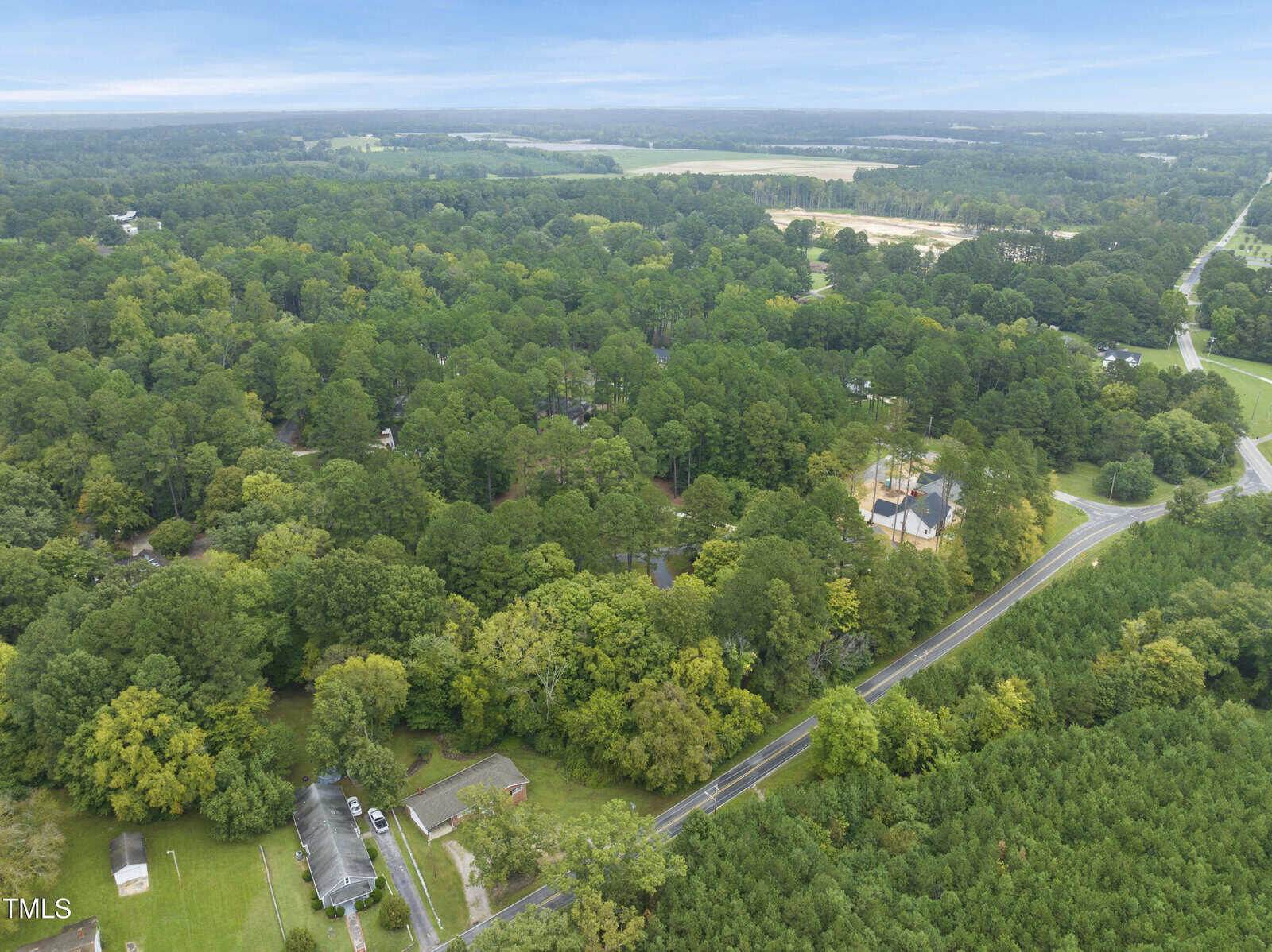 Aerial view of a rural area with dense green trees, a few houses, and winding roads surrounded by forest and open fields under a cloudy sky.