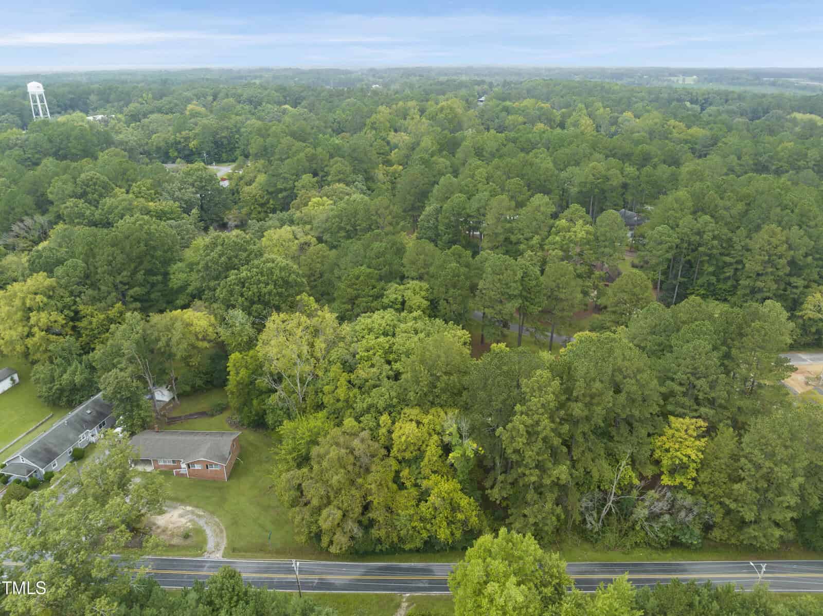 Aerial view of a rural area with dense green trees, a single-story brick house, a road in the foreground, and a water tower visible in the background under a cloudy sky.