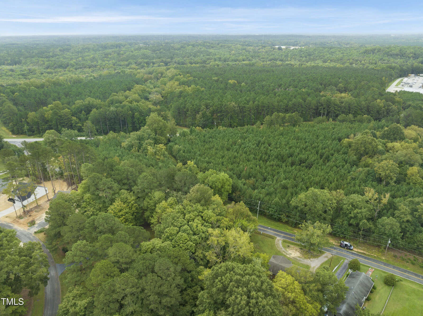 Aerial view of a green, forested area with dense trees, intersected by a road. There are scattered buildings near the bottom and left, and an industrial or commercial site in the upper right corner. The sky is mostly cloudy.