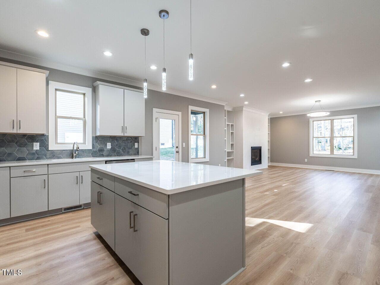 Modern open-concept kitchen and living area with light wood flooring, white cabinetry, a large kitchen island, pendant lighting, and built-in shelving next to a fireplace. Large windows provide natural light.