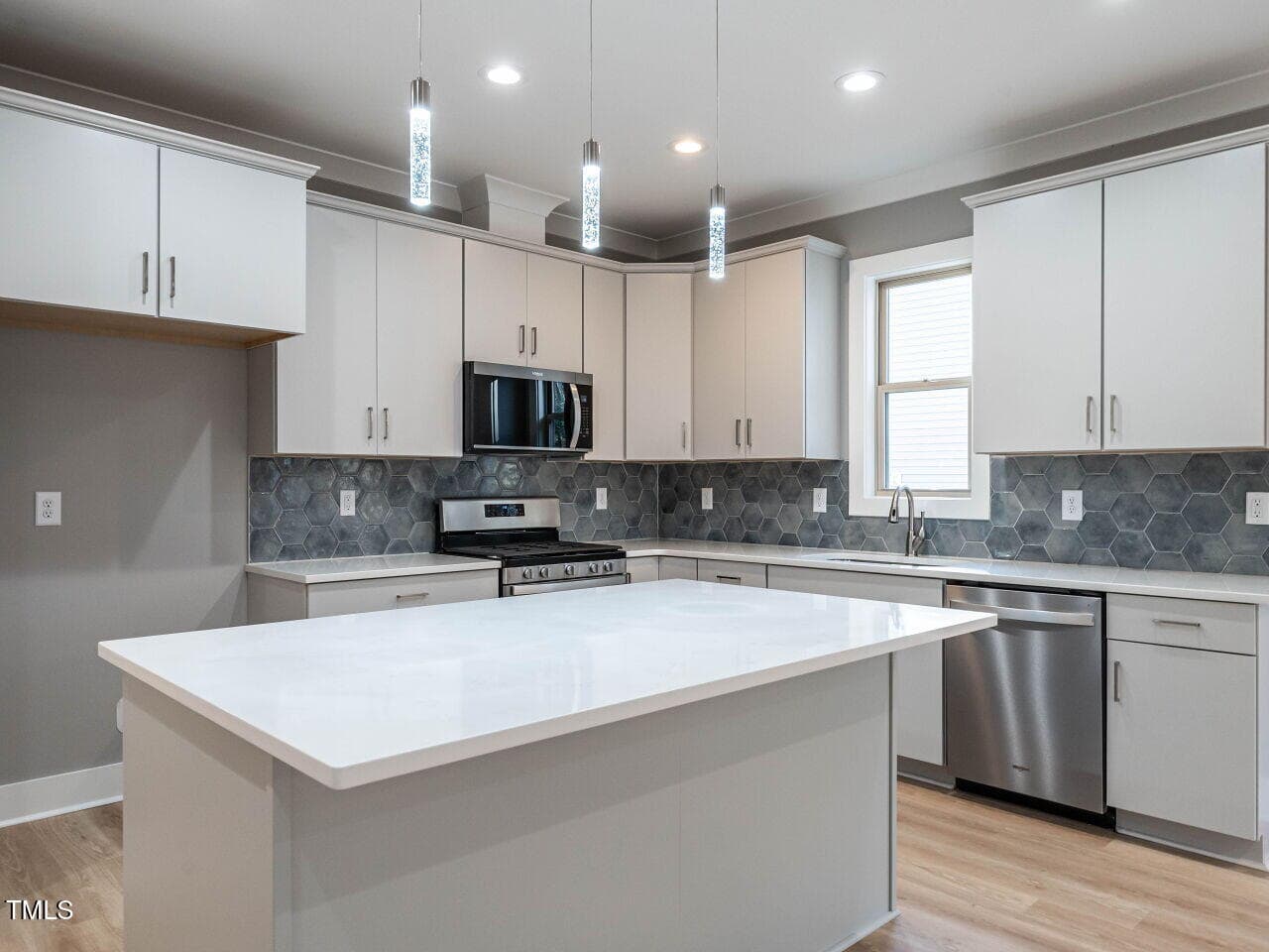 A modern kitchen with white cabinets, stainless steel appliances, a large white island, gray hexagonal tile backsplash, wood flooring, and three pendant lights hanging above the island.
