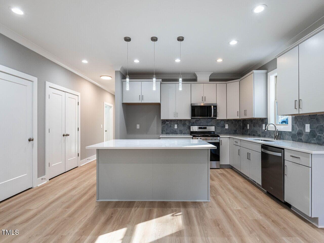 Modern kitchen with light wood floors, a large white island, white cabinetry, stainless steel appliances, hexagonal tile backsplash, pendant lighting, and recessed ceiling lights. Two closed doors are seen on the left wall.