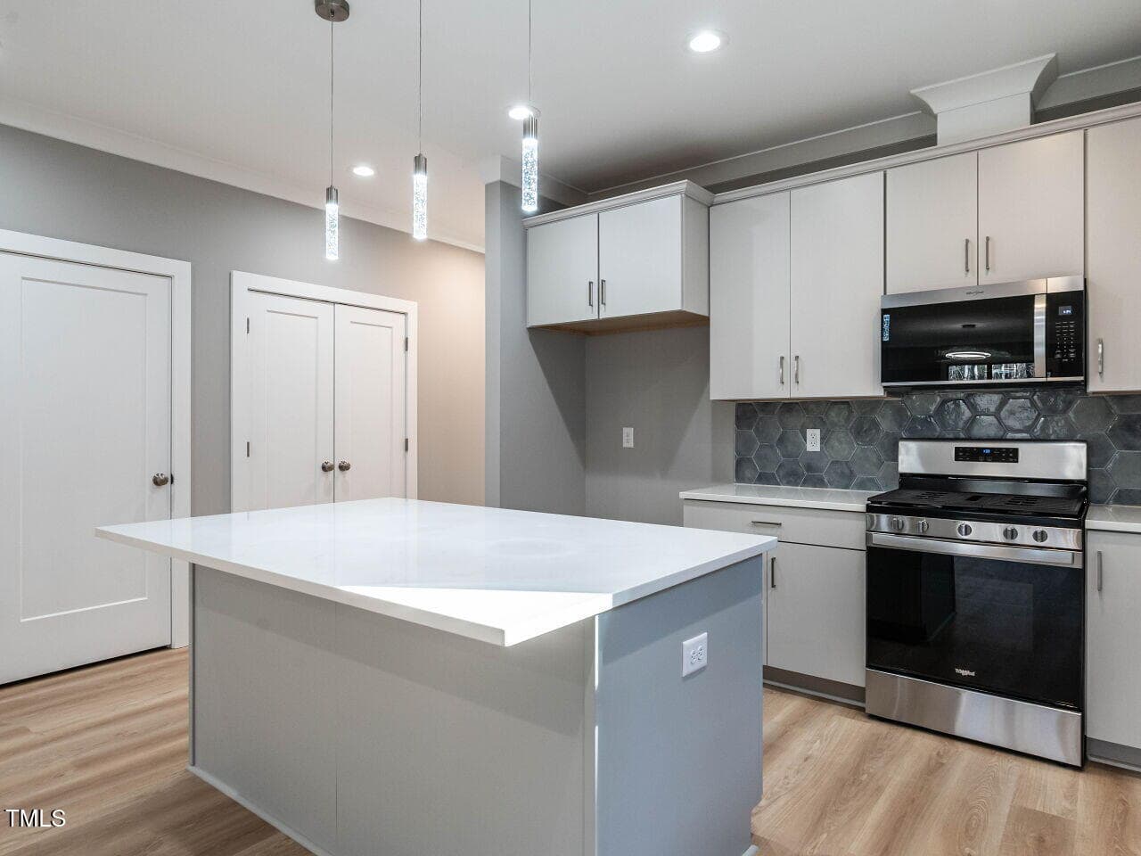 A modern kitchen with light gray cabinets, a white island countertop, stainless steel gas stove, built-in microwave, hexagonal tile backsplash, pendant lights, and light wood flooring. Two closed doors are visible.