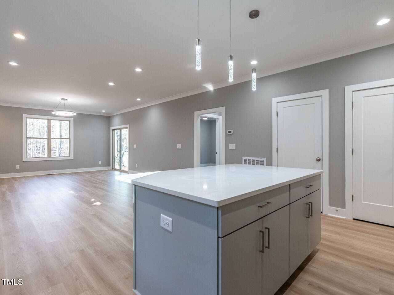 A modern, unfurnished open-concept living space with light wood flooring, gray walls, recessed lighting, and a white kitchen island with cabinets and pendant lights. Doors and large windows are visible in the background.