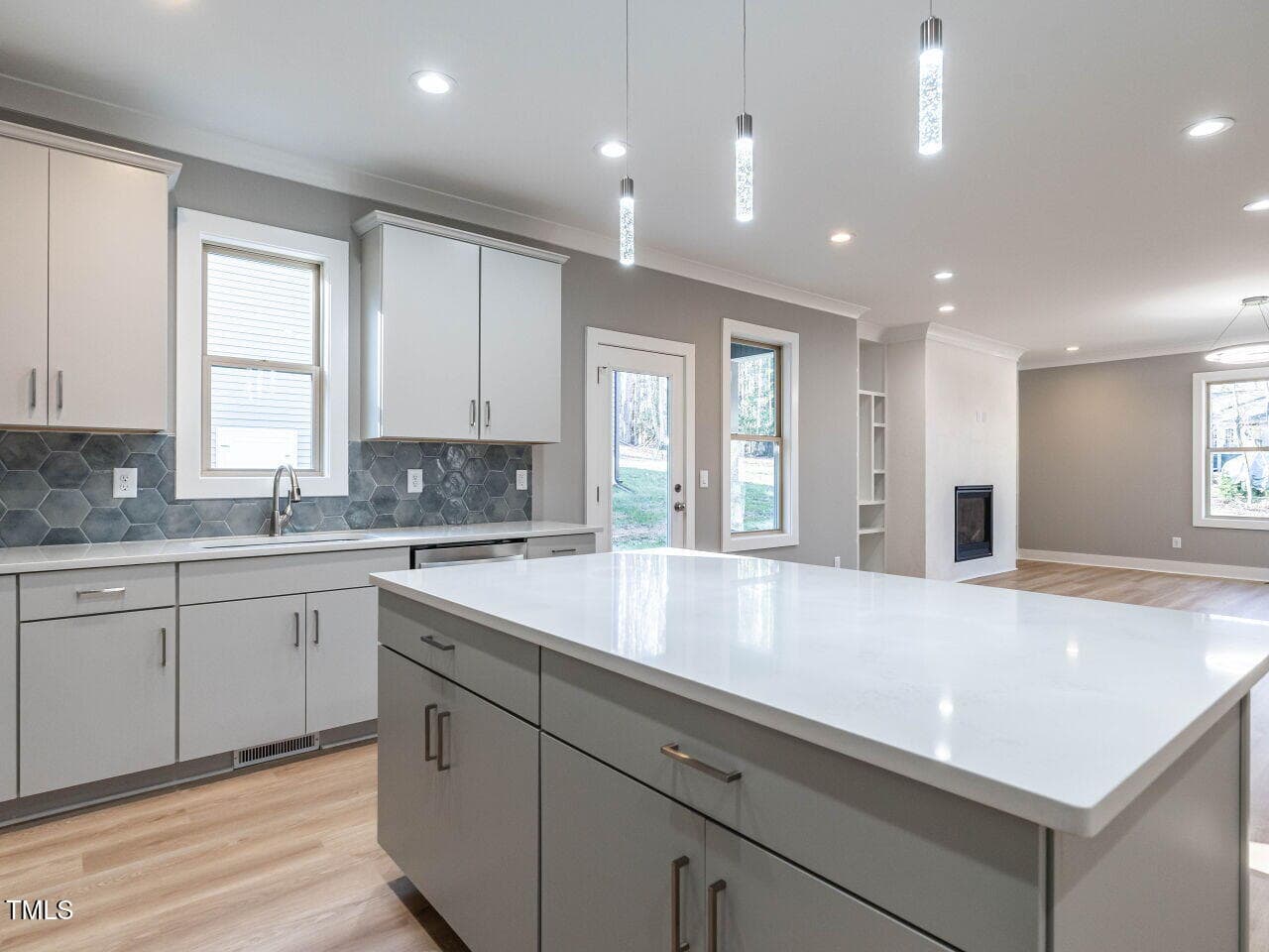 Modern kitchen with white cabinets and drawers, a large island with a white countertop, hexagonal tile backsplash, stainless steel handles, pendant lights, and an open view to a living area with a fireplace.