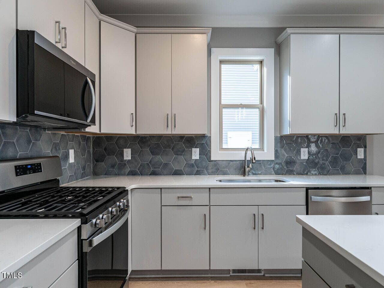 Modern kitchen with light gray cabinets, stainless steel appliances, a gas stove, a microwave, and a dishwasher. Hexagonal gray tile backsplash and a single window above the sink provide natural light.