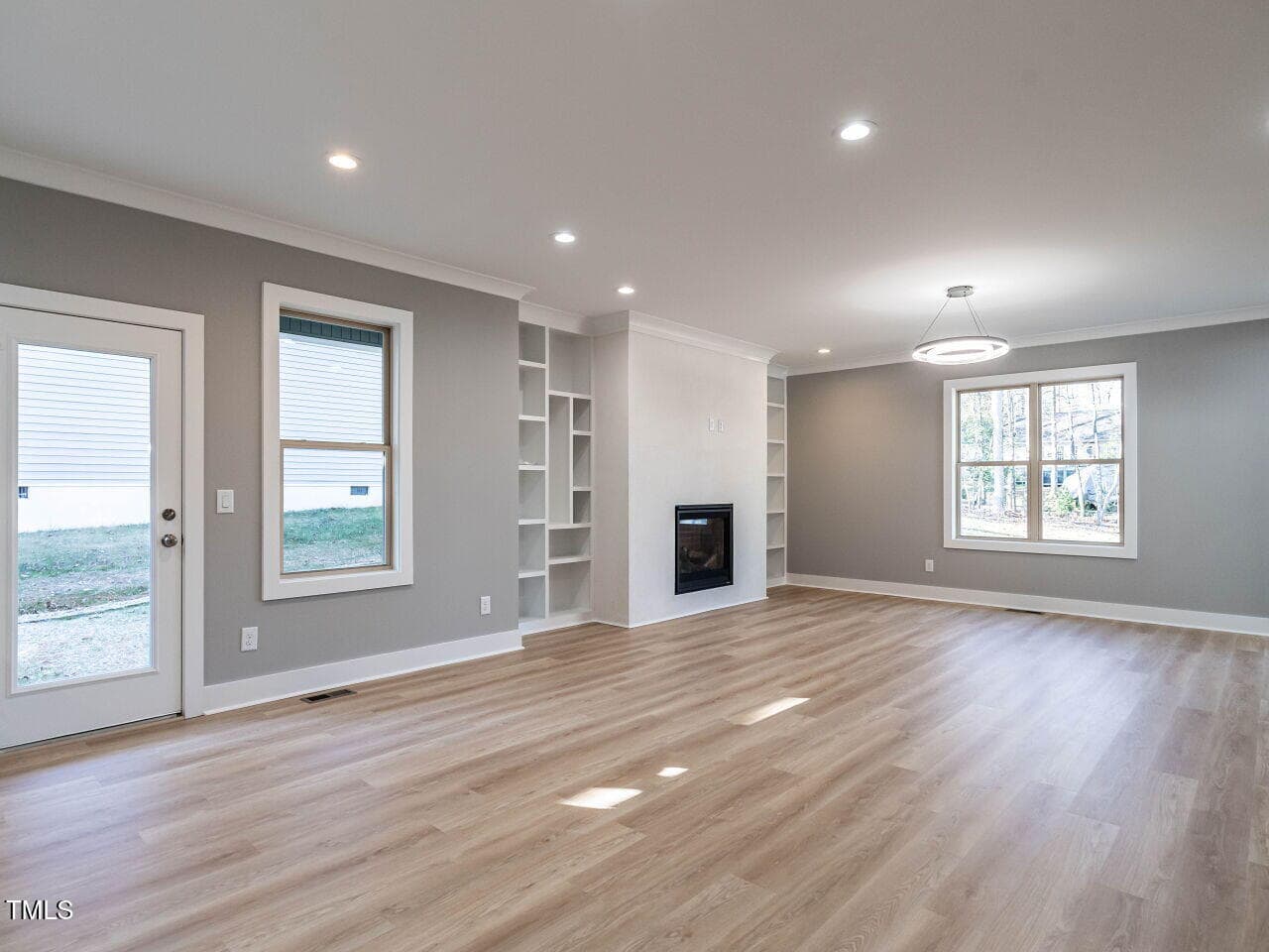 A spacious, empty living room with light wood flooring, gray walls, built-in shelves, a fireplace, and large windows. Recessed ceiling lights and a modern ceiling fixture provide lighting. A glass door leads outside.