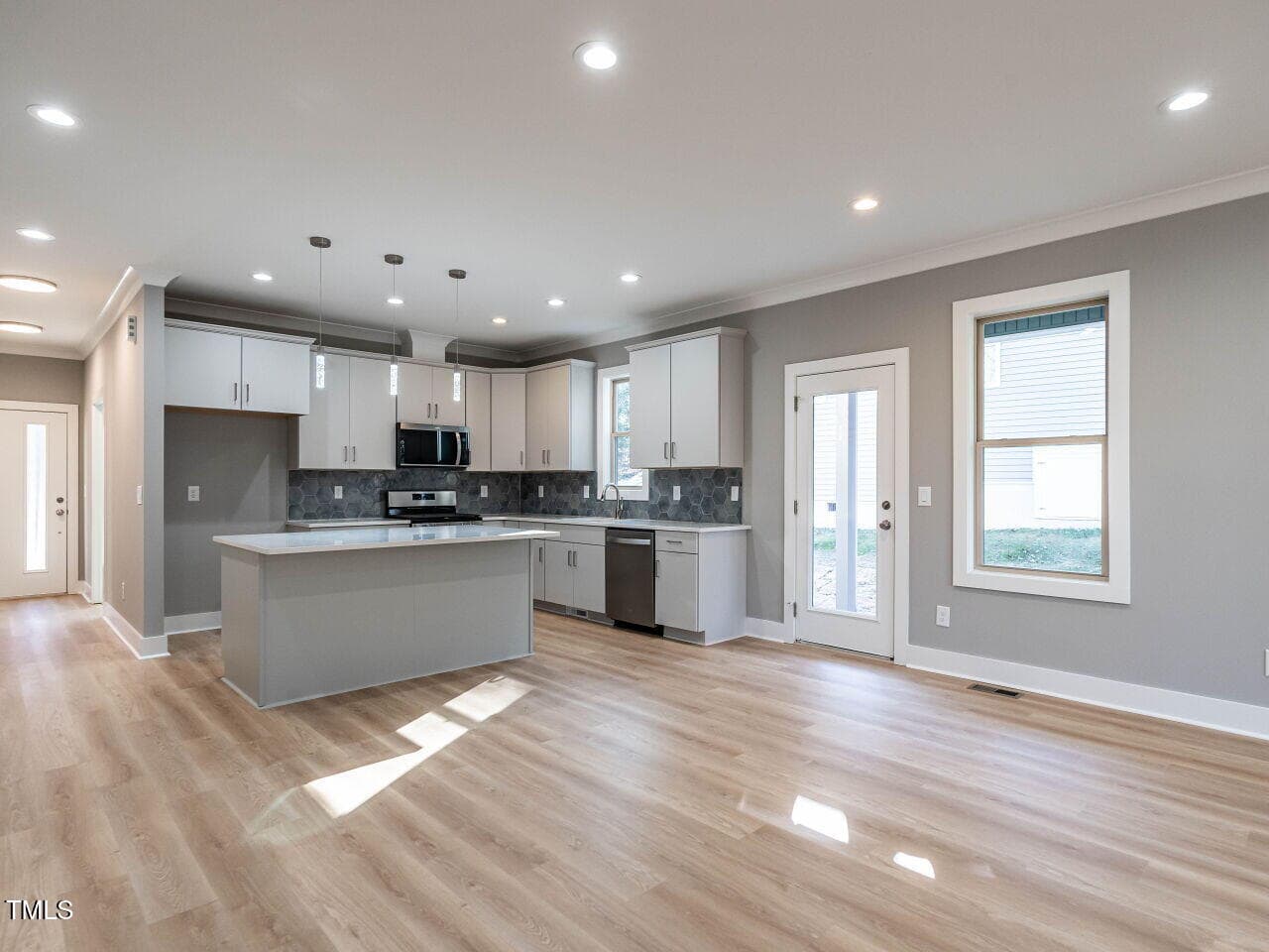 Modern kitchen with white cabinets, gray island, stainless steel appliances, light wood flooring, recessed lighting, and a door with glass panes leading outside. Windows provide natural light to the open space.