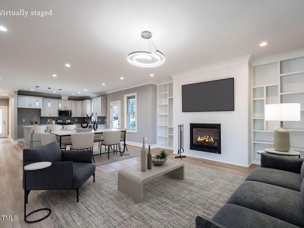 A modern, virtually staged living room with a gray sofa, armchair, coffee table, built-in shelves, and a wall-mounted TV above a fireplace. The open space connects to a kitchen with white cabinets and a dining area.