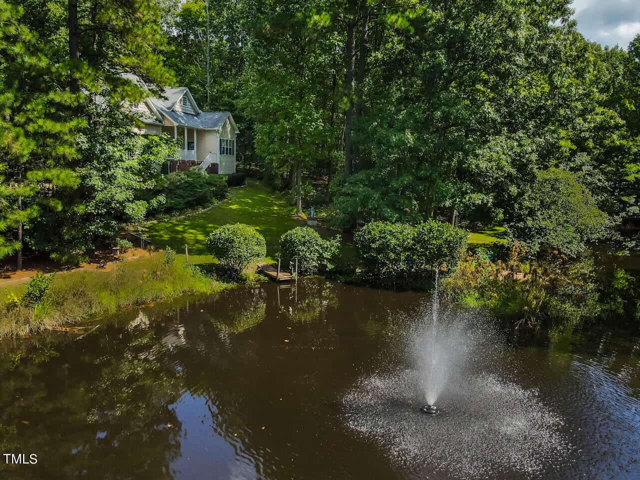 A serene view of a house surrounded by tall trees near a pond. The water is calm, and a fountain sprays in the foreground. Lush greenery and shrubs line the edge of the water, creating a peaceful, nature-filled setting.