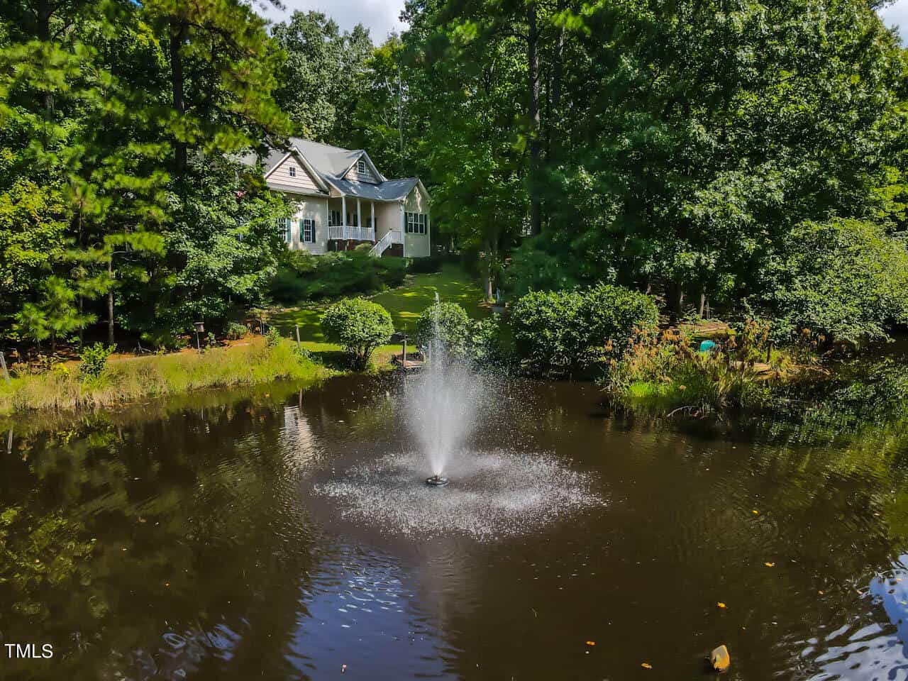 A serene landscape with a house surrounded by dense trees. In the foreground, a small pond features a fountain, creating ripples on the water surface.