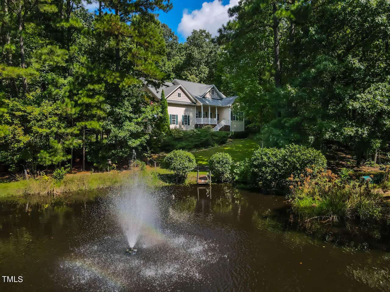 Nestled on 4102 Geneva Drive in Franklinton, this charming cottage-style home is surrounded by lush trees, with a well-maintained garden leading to a small pond. A fountain in the pond creates a spray of water, capturing rainbows under a bright blue sky with scattered clouds.
