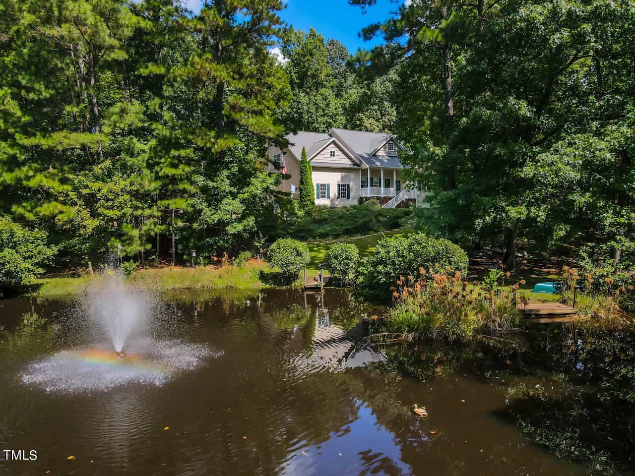 A scenic view of a house surrounded by lush trees, with a peaceful pond in the foreground. The pond features a small fountain creating ripples and a rainbow effect on the water. A dock with chairs is nearby, enhancing the tranquil setting.
