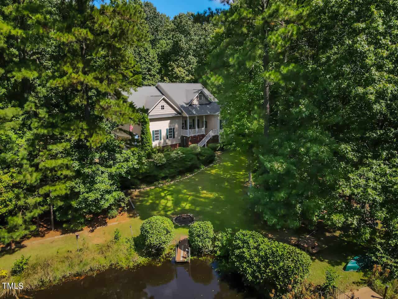 A house surrounded by lush green trees sits near a small pond under a clear blue sky. A wooden dock extends into the water, and the grassy lawn is well-maintained, creating a peaceful, natural setting.