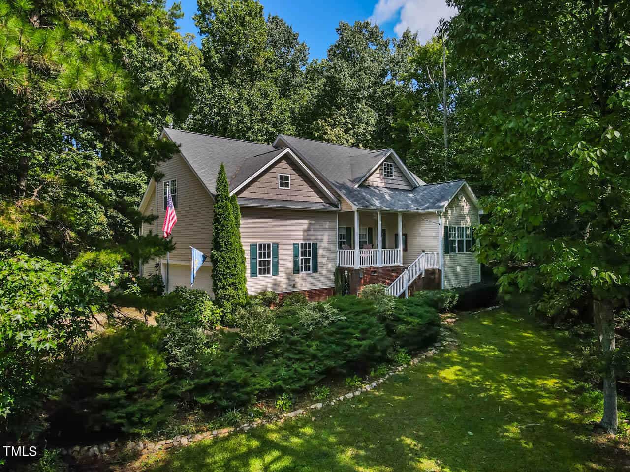 A beige house with a gray roof is surrounded by lush greenery and trees. It features a porch with white railings and steps leading to the entrance. An American flag is displayed on the porch. The sky is blue with scattered clouds.