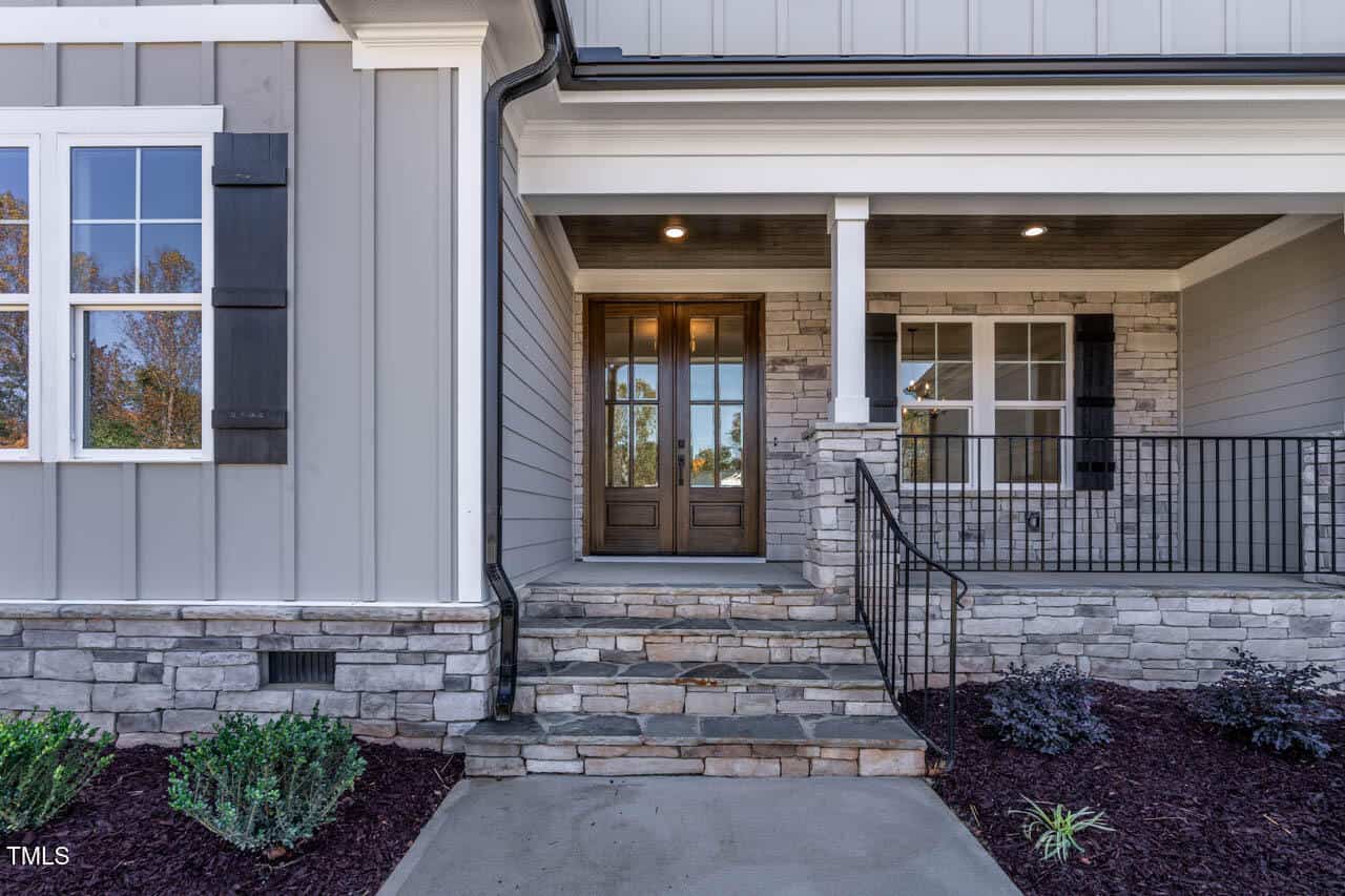 The image shows the front entrance of a house with a wooden double door. The exterior features gray siding and stone accents. There are steps leading up to the door and a black metal railing. The garden is landscaped with small shrubs.
