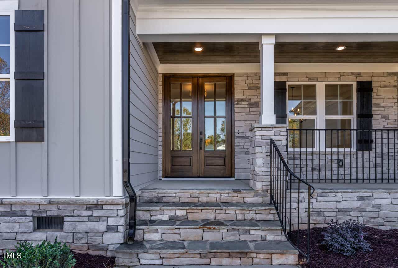 A front view of a house entrance featuring double wooden doors with glass panels, surrounded by stone and horizontal siding. A small staircase leads to the door, with a metal railing on the right. A window is visible on the right side.