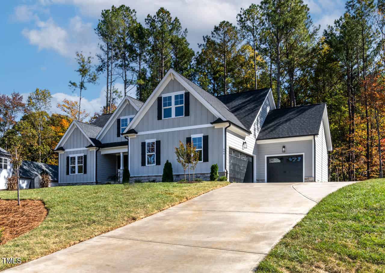 A modern, two-story house with a gray exterior and dark roof is situated on a sloped driveway. The home is surrounded by trees and a well-manicured lawn. There are multiple windows and a garage on the right side.