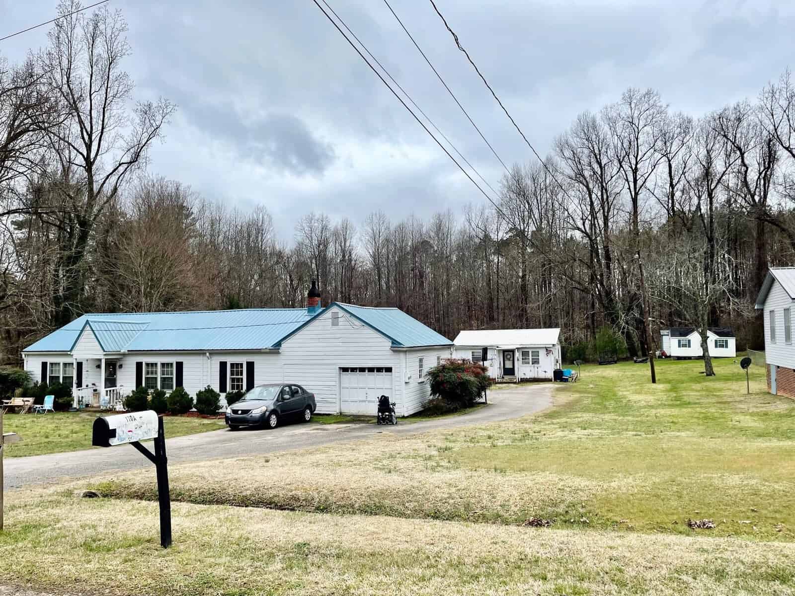 A suburban scene featuring a single-story house with a blue metal roof and an attached garage. A car is parked in the driveway. The yard is expansive with bare trees in the background and a mailbox in the foreground. A smaller building is visible behind the main house.