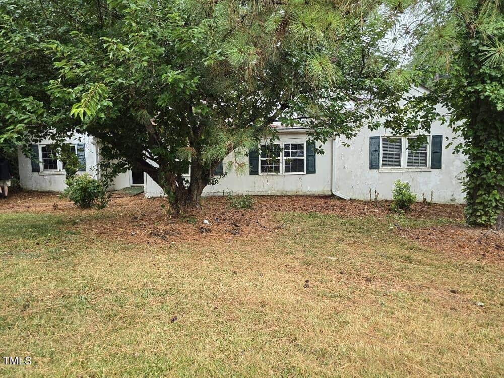 A white, single-story house at 115 Red Bud Drive with dark green shutters partially obscured by trees and overgrown foliage. The lawn surrounding the house appears unkempt with patches of dry grass. The simple, suburban style home in Henderson retains a quiet charm despite its neglected state.