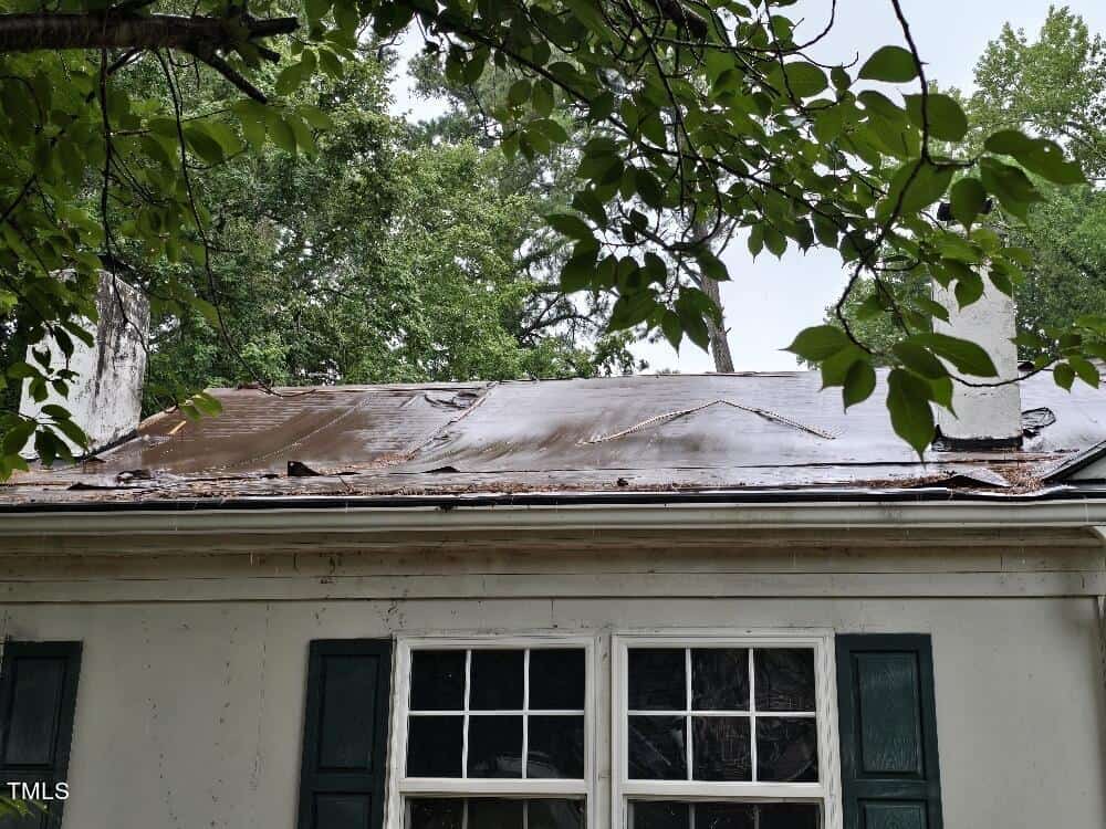 A house with white exterior walls and dark green shutters is pictured. The roof is visibly damaged with rust and wear. Overhanging tree branches partially obscure the view of the roof. The surrounding area appears lush and green.