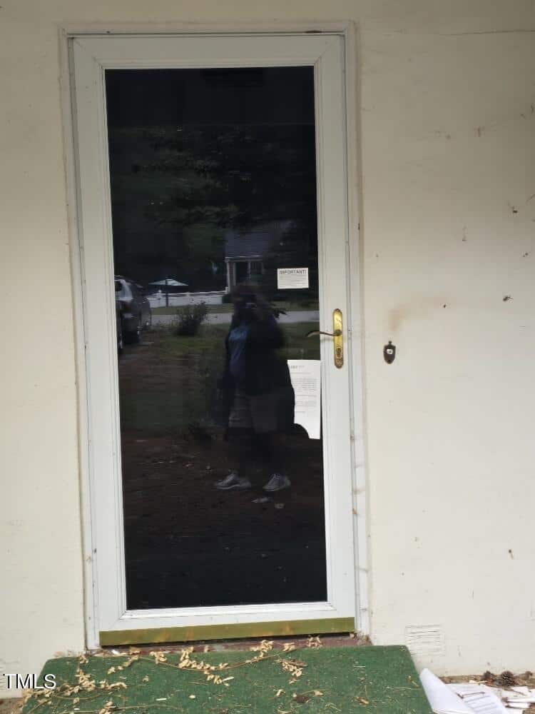 A closed white door with a glass panel is set in a light-colored exterior wall. The reflection of a person taking a photo is visible in the glass. The ground in front has green artificial turf with scattered leaves. A house and vehicle can be seen in the background.