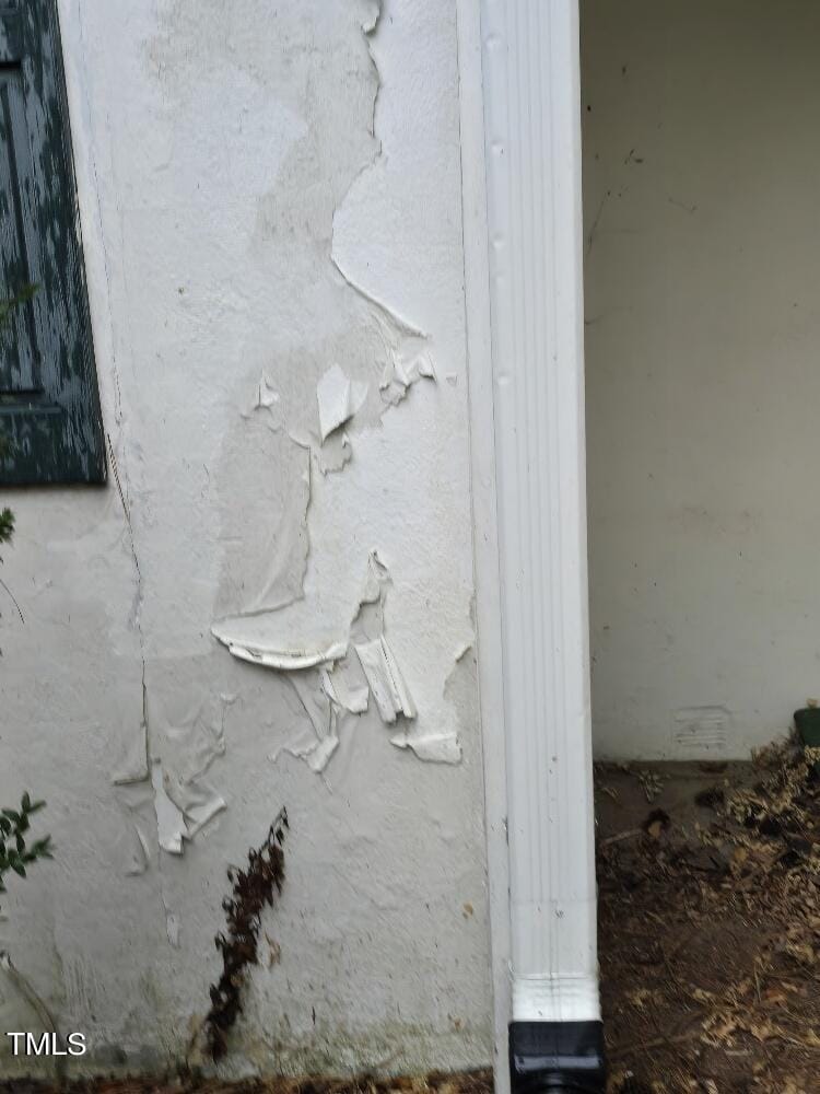 A close-up of the corner of a house's exterior, showing peeling white paint on the walls. There is a white downspout attached to the corner, and some green trim is visible on the left side. Some dark plants and debris are at the base of the wall.
