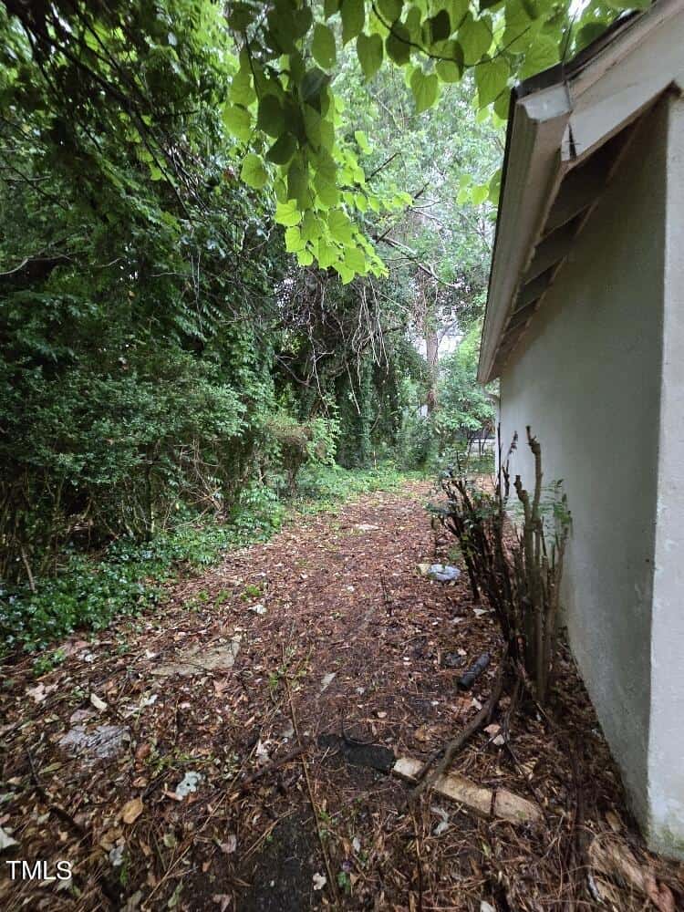 A narrow dirt path runs alongside a house with white exterior walls. Dense green foliage and trees overshadow the path, creating a lush, rustic environment. The ground is covered with fallen leaves and small stone pieces. Overcast light filters through the leaves.