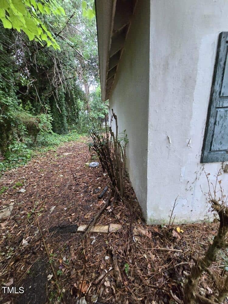 A narrow, overgrown pathway runs alongside a white building with peeling paint and a partially shuttered window. Dense foliage and trees surround the path, creating a secluded, somewhat neglected atmosphere.