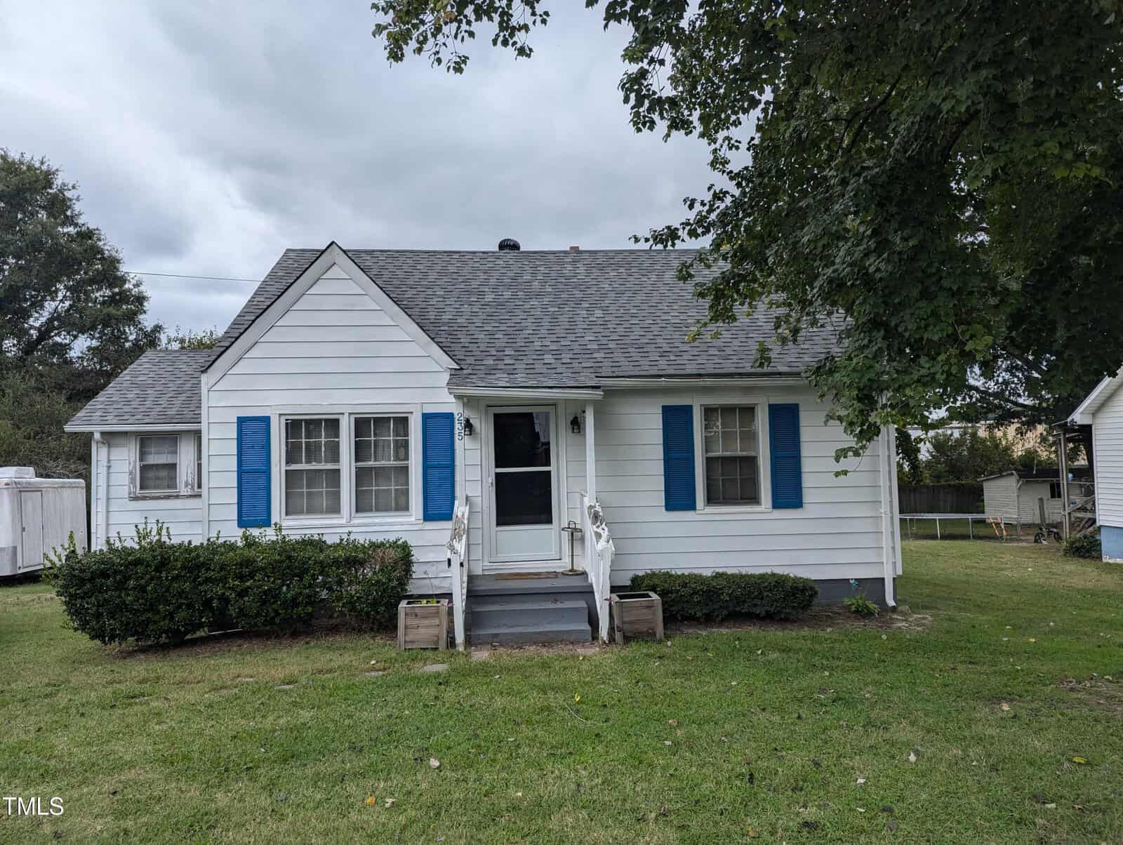 A small white house with blue shutters, a gray shingled roof, and a front porch with steps and handrails sits at 235 Orville Street in Henderson. The house is surrounded by a grassy lawn and bushes, with a few large trees nearby. The sky is overcast.