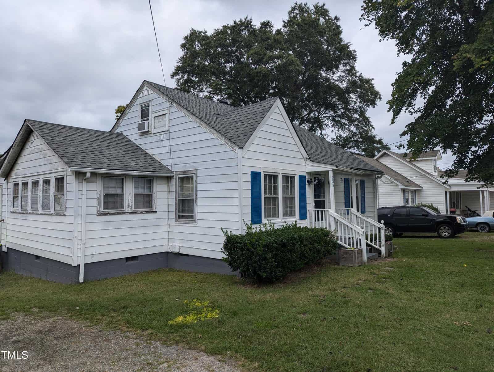 A small white house with blue shutters, featuring a gabled roof and a small porch with steps leading to the entrance. The house is surrounded by grass, and a black car is parked on the gravel driveway to the right. Trees are visible in the background.