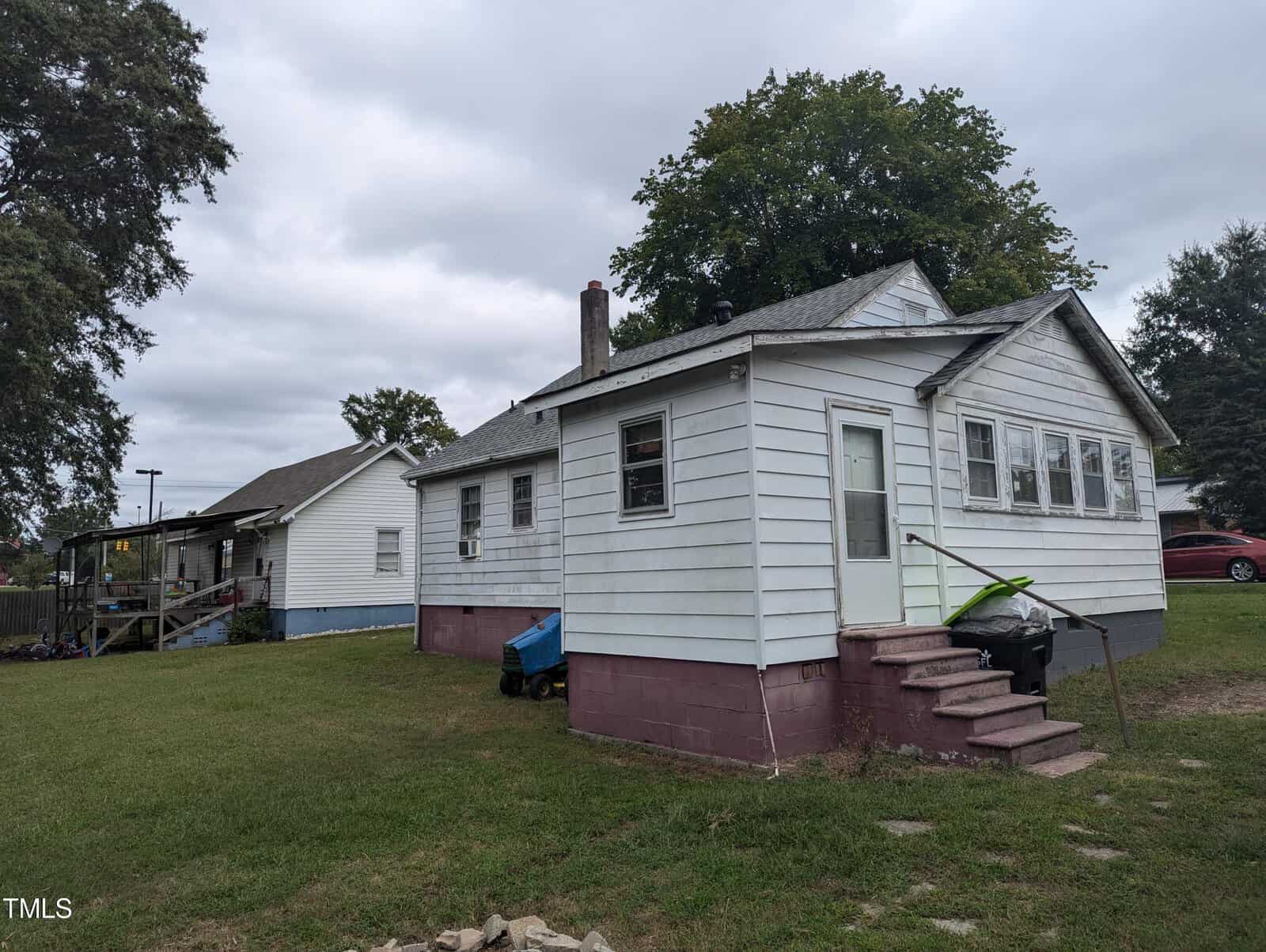 A small white house with a red foundation sits on a grassy yard. The house has a set of steps leading to the side door. There is a blue wheelbarrow and a grill next to the steps. Another house is visible in the background under a cloudy sky.