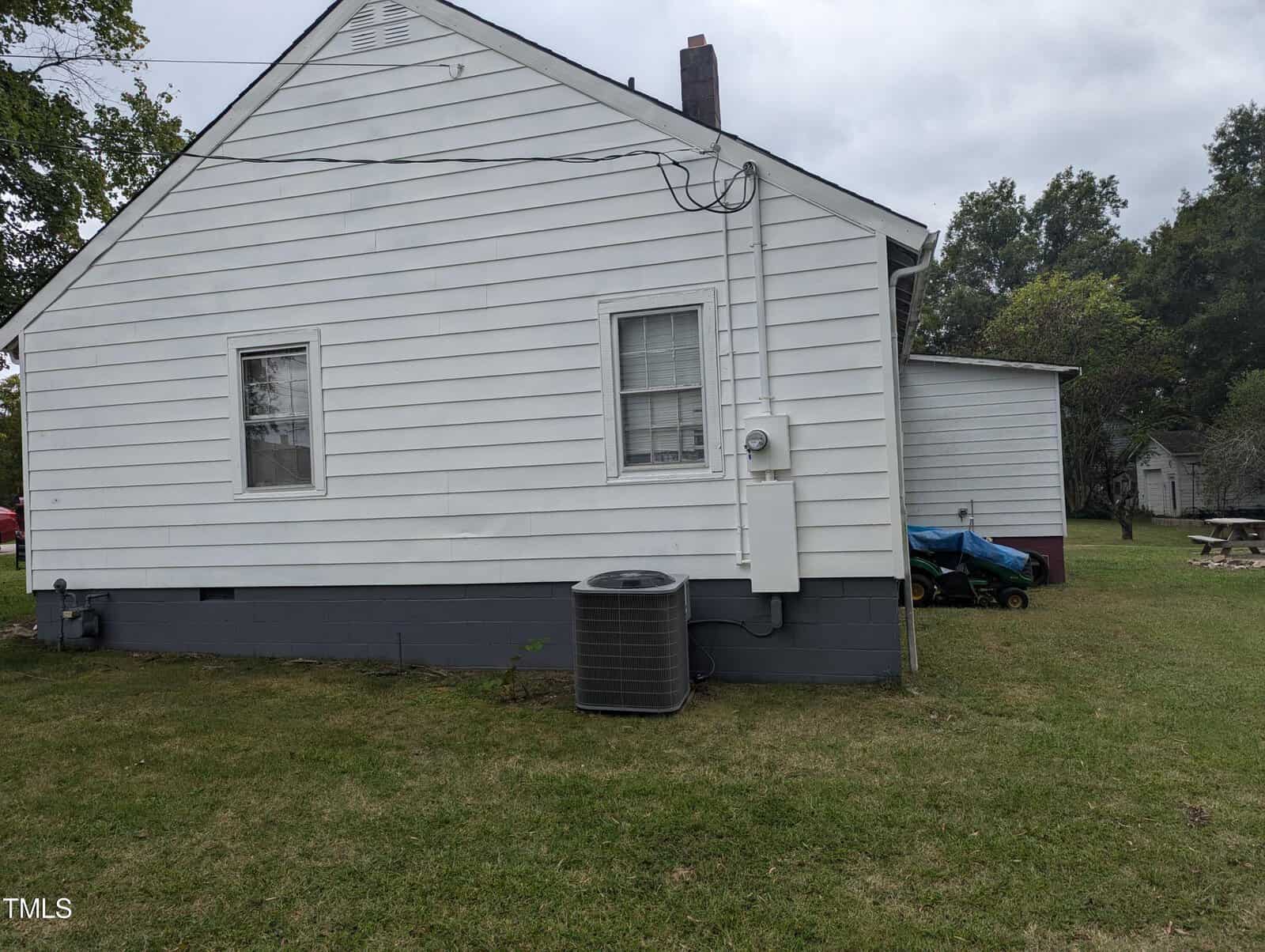 A small white house with horizontal siding and two windows on the visible side. An air conditioning unit is situated against the house, and a wire leads from it to the house wall. There is a grassy yard with trees and a picnic table. The sky is overcast.