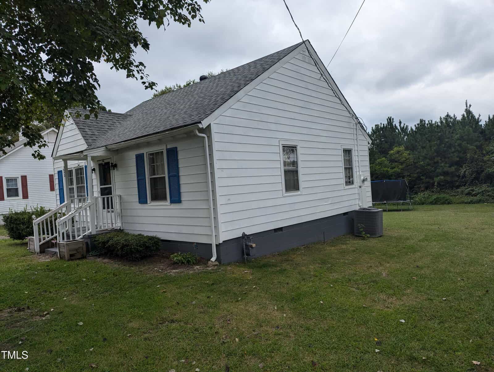 A small white house with blue shutters stands in a grassy yard. The house features a short front porch with white railings and steps. A large tree is partially visible to the left, and a trampoline is seen near the tree line on the right. The sky is cloudy.